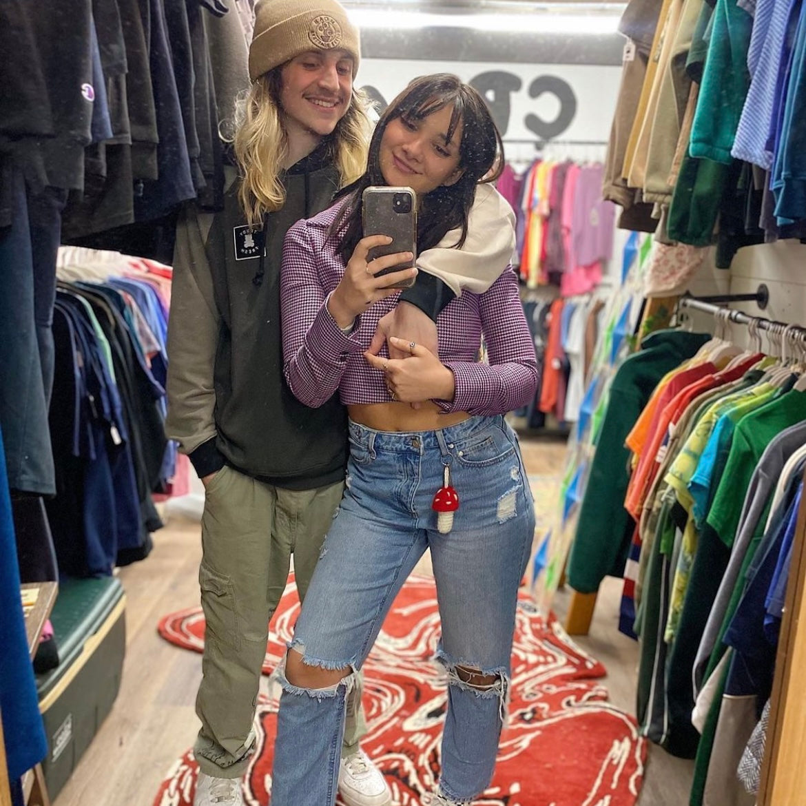 girl and guy taking a mirror selfie. girl has a red and white crochet mushroom keychain hanging from her belt loop